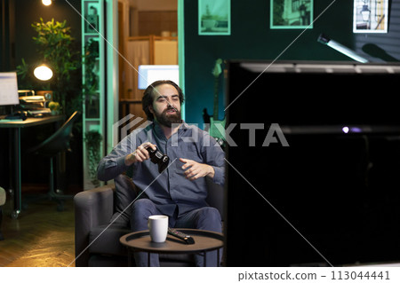 Happy man playing shooter videogame in neon illuminated home, holding controller. Gamer excited to participate in online tournament, battling other players using professional joypad 113044441