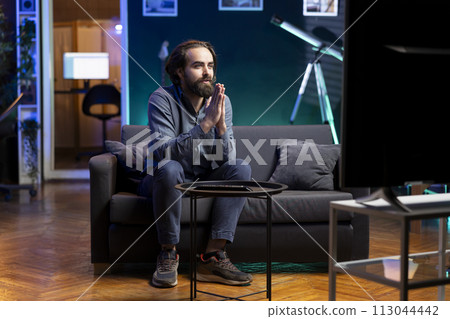 Hopeful man praying for favorite team to win while watching live sports event broadcasted on TV. Supporter sitting on couch at home doing imploring hand gesture, enjoying football competition 113044442