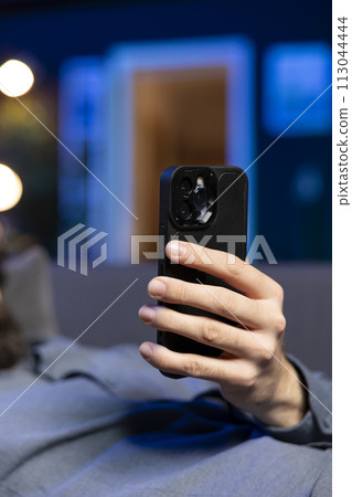 Focus on cellphone used by man in blurry background relaxing at home lounged on couch, texting friends. Close up shot of phone used by person laying down, chatting with mates online Focus on cellphone used by man in blurry background relaxing at home lounged on couch, texting friends. Close up shot of phone used by person laying down, chatting with mates online 113044444