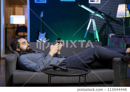 Chilled man relaxing at home lounged on couch, texting friends on smartphone. Person laying down, taking break to chat with mates online using mobile phone in living room 113044446