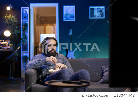 Person using TV to binge series on subscription based streaming services, enjoying bowl of popcorn. Man relaxing in apartment watching VOD shows on television display, eating snack Person using TV to binge series on subscription based streaming services, enjoying bowl of popcorn. Man relaxing in apartment watching VOD shows on television display, eating snack 113044448