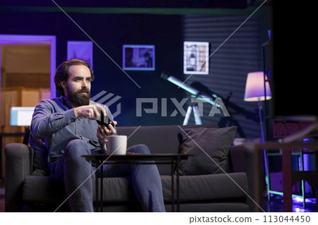 Gamer immersed in virtual world, holding controller in modern apartment, playing videogames. Cheerful caucasian man enjoys gaming session from the comfort of his living room Gamer immersed in virtual world, holding controller in modern apartment, playing videogames. Cheerful caucasian man enjoys gaming session from the comfort of his living room 113044450
