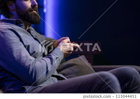 Gamer sitting on couch engaging in online competitive multiplayer tourney using gaming console and gamepad. Player engaging in fun videogames, enjoying leisure time while defeating enemies Gamer sitting on couch engaging in online competitive multiplayer tourney using gaming console and gamepad. Player engaging in fun videogames, enjoying leisure time while defeating enemies 113044451