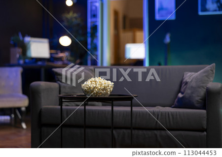 Focus on bowl of popcorn on table with neon illuminated home theater apartment in blurry background. Salty snack prepared for upcoming movie night in cozy RGB lit living room 113044453
