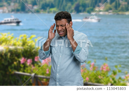 Stressed young man suffering from terrible headache standing outdoors. Man with headache on sea background. Stressed young man suffering from terrible headache standing outdoors. Man with headache on sea background. 113045803