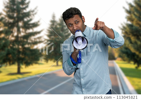 Young indian man talking into megaphone and pointing with index finger. Man with loudspeaker standing outdoors. 113045821