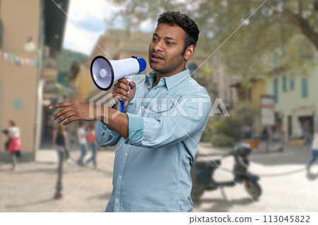 Young man with megaphone standing on blur city street background. Indian man with loudspeaker outdoors. 113045822