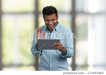 Smiling young man waving with hand while doing video call through digital tablet. Communication via internet. Smiling young man waving with hand while doing video call through digital tablet. Communication via internet. 113045860
