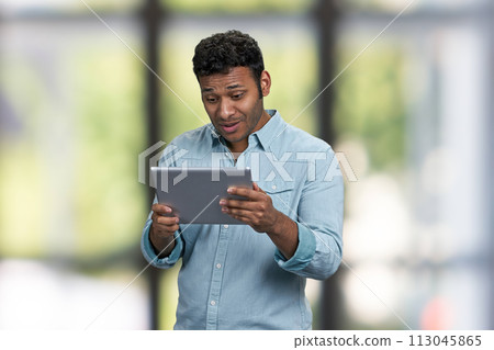 Surprised young man holding digital tablet on interior blur background. People, modern technology and lifestyle concept. Surprised young man holding digital tablet on interior blur background. People, modern technology and lifestyle concept. 113045865