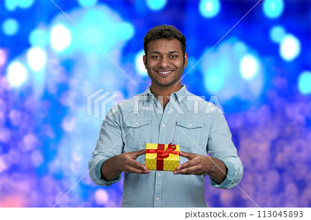 Attractive smiling young man handing gift box to camera. Blue bokeh lights background. Christmas present delivery in time. 113045893