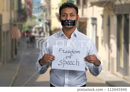 Young man with taped mouth holding paper card with inscription Freedom of speech. Blur narrow city street in the background. Young man with taped mouth holding paper card with inscription Freedom of speech. Blur narrow city street in the background. 113045940