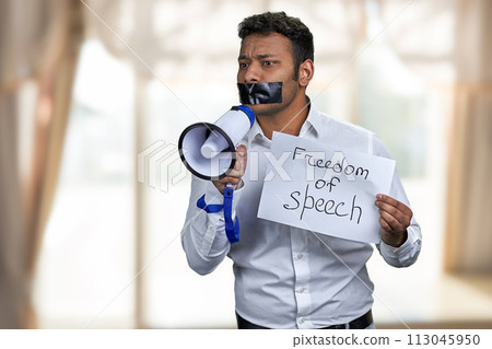 Young man with taped mouth trying to speak into megaphone. Freedom of speech concept. Young man with taped mouth trying to speak into megaphone. Freedom of speech concept. 113045950