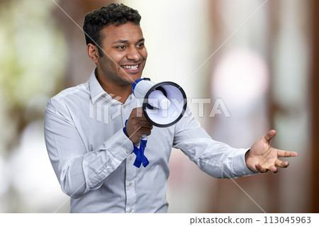Cheerful young man in white shirt talking into megaphone. Abstract bokeh background. 113045963