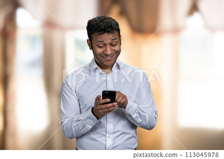Handsome young man in white shirt surfing internet on mobile phone. Cheerful indian man using cell phone on blur interior background. Handsome young man in white shirt surfing internet on mobile phone. Cheerful indian man using cell phone on blur interior background. 113045978