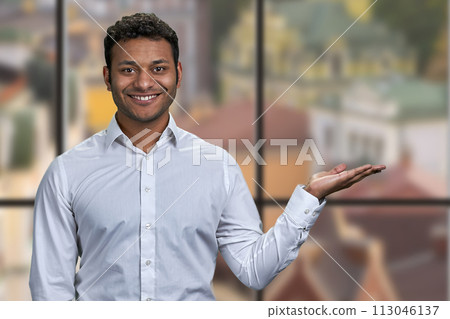 Handsome man in white shirt presenting something with open hand palm. Blur window background with view to city buildings. Handsome man in white shirt presenting something with open hand palm. Blur window background with view to city buildings. 113046137