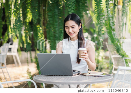 Happy freelancer works on her laptop while holding a coffee cup, sitting at a garden table surrounded by verdant plants. Happy freelancer works on her laptop while holding a coffee cup, sitting at a garden table surrounded by verdant plants. 113046840