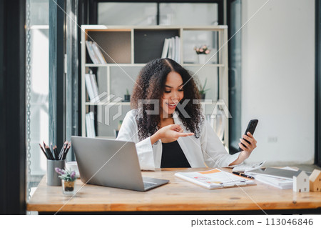 Businesswoman using her smartphone while reviewing documents, with a laptop open on the desk in a well-lit modern office. Businesswoman using her smartphone while reviewing documents, with a laptop open on the desk in a well-lit modern office. 113046846
