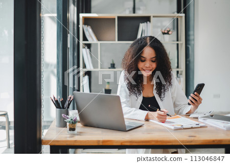 Businesswoman using her smartphone while reviewing documents, with a laptop open on the desk in a well-lit modern office. Businesswoman using her smartphone while reviewing documents, with a laptop open on the desk in a well-lit modern office. 113046847