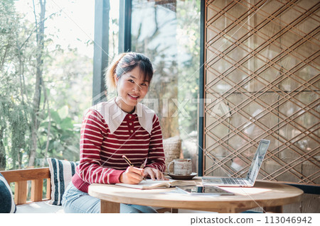 Diligent woman engrossed in writing notes in a notebook with her laptop open on a sunny cafe table. 113046942