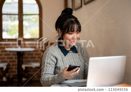 A smiling, positive young Asian woman is working remotely at a coffee shop, working on her laptop. A smiling, positive young Asian woman is working remotely at a coffee shop, working on her laptop. 113049556