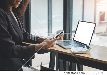 Close-up back view of a business woman working in the office using smartphone and laptop with blank screen, looking at the screen. 113051199