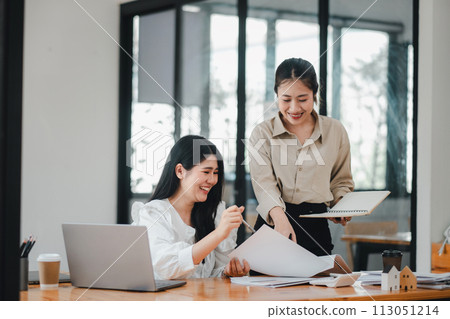 Two professional women share a cheerful moment while collaborating on a project in a bright and modern office environment. 113051214