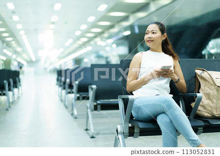 Happy beautiful Asian woman passenger sitting in airport departure terminal alone and sends a message or chats with her friends. Solo female traveler waiting for boarding at the gate in airport. Happy beautiful Asian woman passenger sitting in airport departure terminal alone and sends a message or chats with her friends. Solo female traveler waiting for boarding at the gate in airport. 113052831