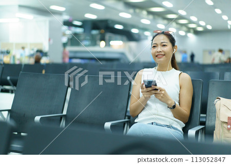 Happy beautiful Asian woman passenger sitting in airport departure terminal alone and sends a message or chats with her friends. Solo female traveler waiting for boarding at the gate in airport. 113052847