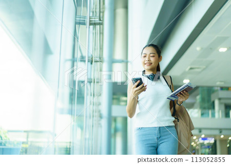 Happy beautiful Asian solo female tourist walking in the airport terminal, woman walking and talking with her friend on mobile phone. Asian woman passenger walks in airport terminal to departure gate. 113052855