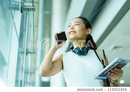 Happy beautiful Asian solo female tourist walking in the airport terminal, woman walking and talking with her friend on mobile phone. Asian woman passenger walks in airport terminal to departure gate. Happy beautiful Asian solo female tourist walking in the airport terminal, woman walking and talking with her friend on mobile phone. Asian woman passenger walks in airport terminal to departure gate. 113052856