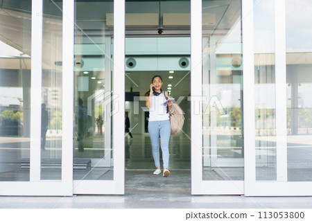 Happy beautiful Asian solo female tourist walking in the airport terminal, woman walking and talking with her friend on mobile phone. Asian woman passenger walks in airport terminal to departure gate. 113053800