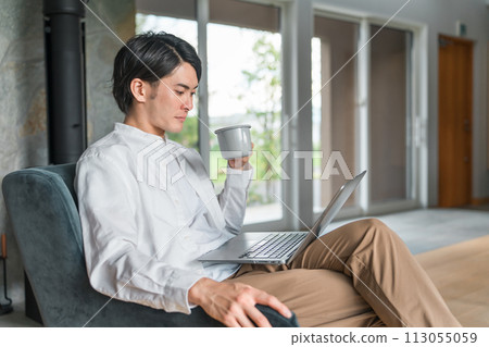 Young Asian man sitting on the sofa in the living room, drinking coffee and looking at a computer 113055059
