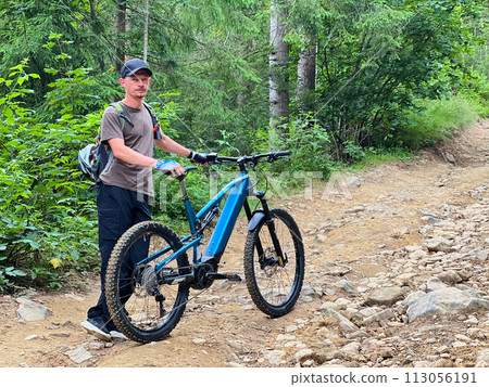 Cyclist man resting on mountain road while riding electric bike outdoors. Portrait of tourist wearing helmet and backpack. Concept of active leisure. 113056191