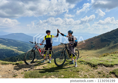 Two cyclists men riding electric bikes outdoors. Male tourists resting on the top of hill, giving high five to each other, enjoying beautiful mountain landscape, wearing helmet and backpack. 113056199
