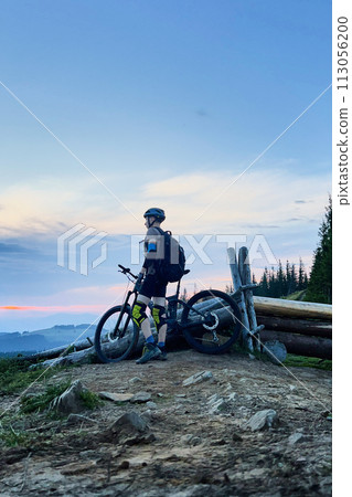 Cyclist man riding electric bike outdoors in the evening. Male tourist resting on hill, enjoying beautiful mountain landscape, wearing helmet and backpack. Concept of active leisure. 113056200