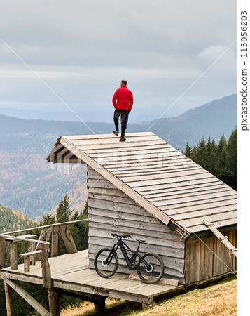 Person resting on edge of steeply sloped wooden rooftop in mountainous area after riding on electric mountain bike on cloudy day. Below the rooftop, there is wooden deck with mountain bicycle. Person resting on edge of steeply sloped wooden rooftop in mountainous area after riding on electric mountain bike on cloudy day. Below the rooftop, there is wooden deck with mountain bicycle. 113056203