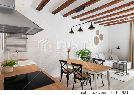 Horizontal shot of a loft-style kitchen-living room with a white wall of wooden beams with modern appliances, a dining table and a sofa corner in a relaxation area in a new apartment Horizontal shot of a loft-style kitchen-living room with a white wall of wooden beams with modern appliances, a dining table and a sofa corner in a relaxation area in a new apartment 113059213