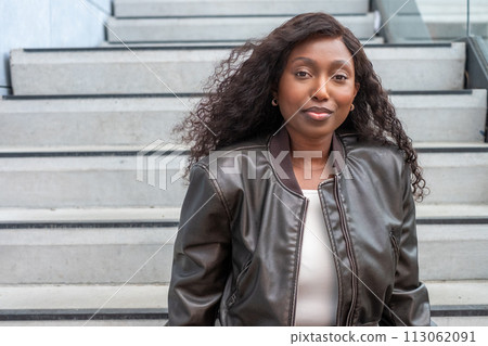 Confident Black Woman in Leather Jacket Seated on Urban Stairs 113062091
