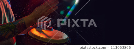 Close-up of male hands, musician playing bongo drums on dark background with stage lights. African culture live event 113063866