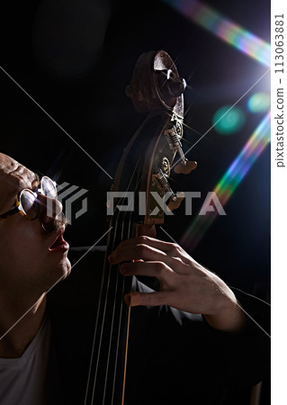 Male musician playing double bass against black background with lights elements. Classical music performance. 113063881
