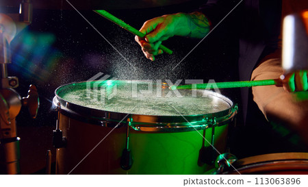 Male hand playing drum with stick on dark background with green hued smoke and light flares. 113063896