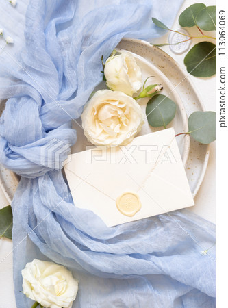 Envelope near light blue fabric knot and cream roses on plates top view copy space, wedding mockup 113064069
