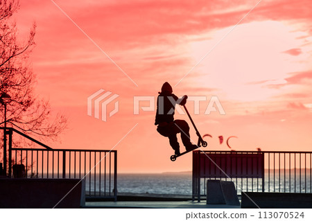 Unrecognizable teenage boy silhouette showing high jump tricks on scooter against 113070524