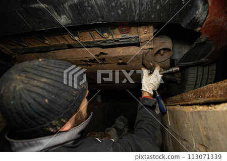 A mechanic unscrews a bolt under a car with a screwdriver to repair a broken part. Repairing a car in a garage in a special pit. Preparing the machine to replace a broken part. A mechanic unscrews a bolt under a car with a screwdriver to repair a broken part. Repairing a car in a garage in a special pit. Preparing the machine to replace a broken part. 113071339