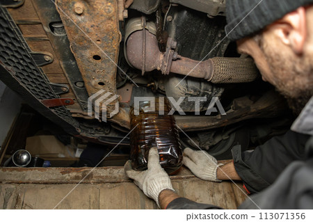 Black motor oil is drained under the car in a pit in the garage. Car maintenance in the garage with engine oil change. The engine oil is drained into a jar to be replaced with new oil. Black motor oil is drained under the car in a pit in the garage. Car maintenance in the garage with engine oil change. The engine oil is drained into a jar to be replaced with new oil. 113071356