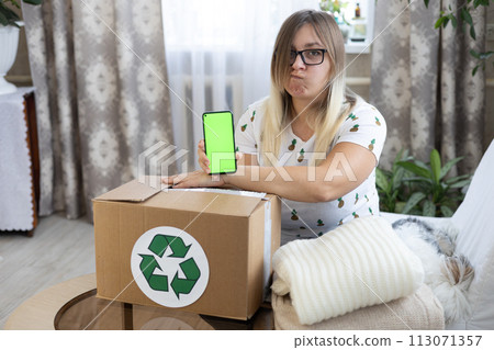 A funny woman with a sad face shows a chrome key phone screen over a box with things. Recycling sign on boxes with things standing on a table in front of a funny woman. A funny woman with a sad face shows a chrome key phone screen over a box with things. Recycling sign on boxes with things standing on a table in front of a funny woman. 113071357