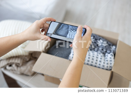 Natural wool sweaters are folded in a box with a recycling sign for reuse. A housewife takes pictures of folded clothes with her phone camera and places an advertisement for sale on the Internet. 113071389