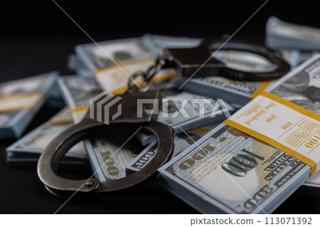 Handcuffs on packs of dollars on a black background. Packs of dollars sealed with bank tape lie with handcuffs symbolizing punishment for theft and bribery. Handcuffs on packs of dollars on a black background. Packs of dollars sealed with bank tape lie with handcuffs symbolizing punishment for theft and bribery. 113071392