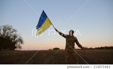 Young male military in uniform jogging with flag of Ukraine on meadow at dusk. Soldier of ukrainian army waving blue-yellow banner in honor of victory against russian aggression. End of war. Slow 113071481
