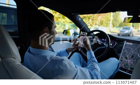Successful businessman browsing modern smartphone while riding an autonomous self driving electric car. Male entrepreneur using phone during riding on electrical auto with autopilot at urban road 113071487